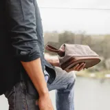Neceser verde de tela canvas combinado con símil cuero marrón. Tiene apertura superior con cierre metálico estructural, doble bolsillo interno (uno con cierre y otro abierto con división) y base con cierre que actúa como bolsillo extra.