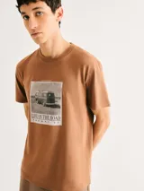 Camiseta blanca de algodón con cuello redondo y mangas cortas. Estampa rectangular con borde negro que contiene la frase "NATURAL ATMOSPHERE" en letras mayúsculas y una imagen de montañas y mar.