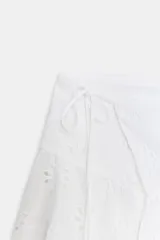 Falda corta blanca de tiro medio con bordados perforados (ojales) y bajo con volantes. Presenta un detalle de lazo ajustable en un lateral de la cintura.