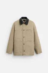 Chaqueta de trabajo color beige con cuello de cuero negro, corte recto, cierre frontal con botones a presión, mangas largas y bolsillos de parche en el pecho y en la parte inferior delantera.