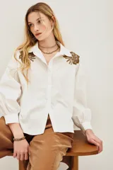 Camisa blanca de algodón con cuello clásico, mangas largas abullonadas con puños anchos y cierre frontal de botones. Presenta aplicaciones bordadas en dorado con detalles de animal print en los hombros.