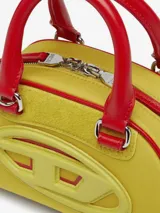 Bolso tipo mini bandolera con forma de domo, color amarillo vibrante con ribetes y asas superiores en rojo. Presenta el logo ovalado 'D' de Diesel en relieve en el frente.