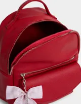 Mochila pequeña de cuero sintético color blanco con bolsillo frontal con solapa y cierre superior. Incluye un charm colgante de moño a rayas rojas y blancas con dije de corazón plateado.