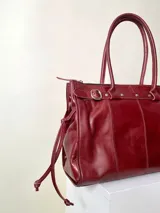 Bolso grande de cuero marrón con textura tipo cocodrilo, estructura rígida y asas para llevar al hombro. Tiene una tira con tachas y hebillas en cada extremo, también lleva lazos en los costados.