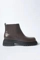 Bota corta tipo Chelsea de cuero marrón oscuro, con elásticos acanalados laterales y puntera cuadrada. Posee una suela track gruesa y dentada de goma negra.