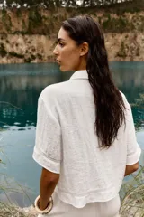 Camisa blanca de cuello solapa y manga corta, con bolsillo delantero y detalle de vainicas a tono. Cierre frontal con botones ocultos por solapa.