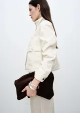 Chaqueta corta color crema, de estilo tejano vaquero, con cuello alto, cierre frontal con botones metálicos, mangas largas con puños abotonados y dos bolsillos de parche con solapa y botón en la parte delantera.