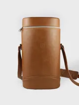 Matera cilíndrica de cuero color camel, con cierre superior y correa ajustable para llevar al hombro. Presenta un escudo grabado en el frente.