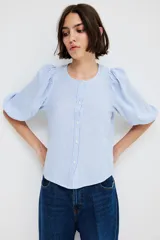 Blusa celeste y blanca a rayas verticales, de corte recto, con cuello redondo, cierre frontal con botones y mangas 3/4 abullonadas con puños elásticos.
