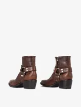 Bota corta de cuero marrón con acabado envejecido, punta estilizada y taco bajo. Presenta un detalle de arnés desmontable con herrajes metálicos plateados alrededor del tobillo.