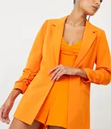 Blazer de sastrería alargado color naranja, con cuello con solapa, manga larga con pliegas en el puño y abertura frontal total. Forro interno de poliéster.