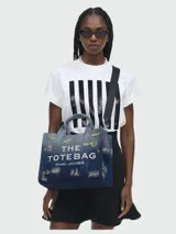 Bolso tote Marc Jacobs de tamaño mediano confeccionado en denim azul con efecto desgastado. Presenta el logo "THE TOTE BAG MARC JACOBS" estampado en blanco en el frente, doble asa corta superior y correa bandolera extraíble de tela negra.