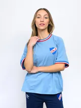 Remera blanca de corte clásico con cuello redondo y mangas cortas con puños tricolor (azul, blanco y rojo). Presenta el logo de Umbro bordado en el pecho izquierdo y el escudo del Club Nacional de Football bordado en el pecho derecho.