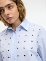 Camisa blanca de algodón con cuello camisero, cierre frontal con botones y tachas plateadas en el frente.