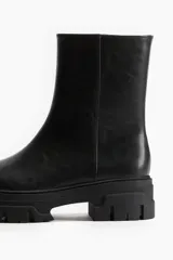 Botas negras de caña media confeccionadas en cuero sintético, con puntera redondeada y cierre lateral con cremallera. Presentan suela track de goma.