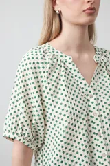 Camisa de viscosa con estampado de pequeños lunares verdes sobre fondo blanco o crema. Presenta escote en V con botones frontales y mangas cortas abullonadas con elástico en el puño.