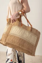 Bolso tipo shopper de rafia color beige con franja horizontal color camel. Tiene asas de cuero color marrón para llevar al hombro y cierre superior.