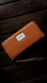 Billetera larga con cierre perimetral, color marrón con textura de cuero granulado. Posee una placa metálica plateada con el logo "Lincoln's Bags & Accessories" en el frente y una correa de mano desmontable.