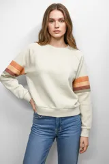 Buzo color crema de corte holgado con mangas raglán y puños ajustados. Presenta tres franjas horizontales en tonos naranja y marrón en la parte superior de ambas mangas.