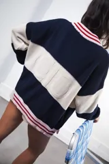 Cardigan de punto grueso con diseño a rayas horizontales en color crema, rojo y marrón. Presenta cuello redondo, cierre frontal con botones oscuros y ribetes a rayas marrones y celestes en cuello, puños y ruedo. Lleva un pequeño bordado con el logo de la marca en el pecho.