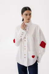 Camisa blanca de algodón con cuello alto con volados, cierre frontal con botones, mangas largas con puños abotonados, bolsillo en el pecho con corazón bordado y parches de crochet en los codos.