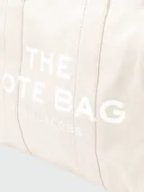Bolso tote negro de lona con asas cortas y estampado frontal en letras blancas con la leyenda "THE TOTE BAG MARC JACOBS".