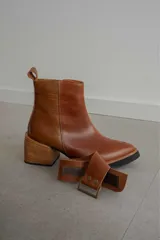 Bota corta de cuero marrón con puntera redondeada y taco cuadrado. Presenta una correa con hebilla alrededor del tobillo y cierre lateral.