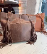 Cartera tipo tote de cuero color marrón con flecos largos decorativos que cuelgan de las asas. Cuenta con un bolsillo externo frontal con costuras a la vista, múltiples compartimentos internos y llavero integrado.