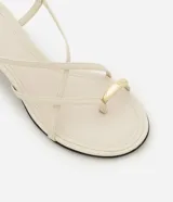 Sandalia femenina confeccionada en material sintético color crema, con punta redonda, tiras finas asimétricas, dedera con detalle bold y hebilla lateral. Prenda con salto mediano fino.