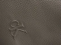 Riñonera de poliuretano color gris topo, con textura granulada y logo 'CK' grabado en el frente. Posee una correa ajustable de tela marrón oscuro.