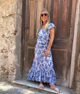 Vestido largo con estampado paisley en tonos azules y violetas sobre fondo blanco. Presenta escote en V, mangas cortas con volados y un ruedo con volado.