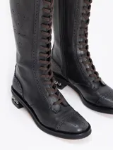 Bota de caña alta color negro, confeccionada en cuero vacuno, con cordones frontales marrones y cierre lateral. Presenta un diseño picado en la capellada y un taco de 4 cm con detalle de esferas metálicas.