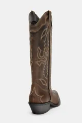 Bota cowboy de caña media, color marrón oscuro con efecto desgastado. Presenta bordados decorativos en hilo claro en el cuerpo y puntera afilada. Posee tacón medio y tiradores laterales para facilitar el calce.