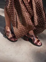 Sandalias bajas de cuero color marrón con tiras anchas cruzadas sobre el empeine y hebilla metálica dorada en forma de estribo.