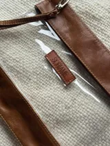 Necessaire de diseño compacto confeccionado en cuero color marrón combinado con material sintético transparente. Presenta cierre superior, costuras reforzadas y una etiqueta frontal de cuero con el logo de la marca grabado.