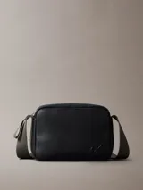 Bolso bandolera rectangular de poliuretano texturizado color negro, con cierre de cremallera superior y logo de Calvin Klein grabado en relieve en la parte frontal inferior. Posee una correa ancha de tela ajustable en tono verde oliva/grisáceo.