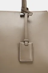 Bolso de mano tipo satchel o tote pequeño, color topo o gris pardo, con doble asa superior y cierre principal por cremallera. Presenta un colgante rectangular de cuero sintético colgando de una de las asas.