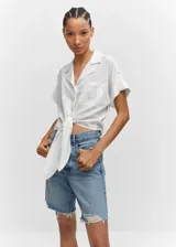 Camisa blanca de lino con cuello camisero, manga corta con vuelta, diseño crop y oversize, bolsillo de parche en la parte delantera, cierre de botones y lazo ajustable.