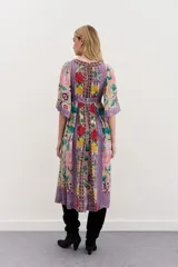 Vestido largo de viscosa con mangas al codo y falda amplia. Presenta un estampado floral vibrante sobre fondo beige, con detalles y bordes en tonos morados y patrones geométricos. Tiene escote en V y cierre trasero.
