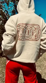 Hoodie color crema con capucha y cordón ajustable. Estampado en la espalda con un paisaje de montañas, árboles y un oso.