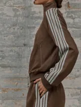 Conjunto deportivo marrón de dos piezas: campera con cremallera, cuello alto, mangas largas con puño y detalles de rayas blancas en mangas y laterales del pantalón. Pantalón recto de cintura alta con cordón ajustable.