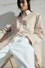 Camisa oversized de sarga color beige con rayas verticales marrones, cuello clásico, cierre frontal con botones ocultos, mangas largas con puños abotonados y bajo recto.