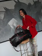 Bolso de viaje tipo weekender de cuero color marrón con detalles en rojo en los cierres. Tiene doble asa corta y correa de hombro ajustable y extraíble. Cuenta con bolsillos internos y externos.