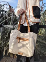 Bolso tipo tote color crema, con detalles en cuero color marrón. Tiene un bolsillo con cierre en la parte superior y un parche de cuero con el logo de la marca en la parte inferior.