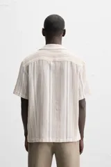 Camisa de manga corta con cuello bowling y corte relaxed fit. Diseño a rayas verticales en tonos beige y blanco. Cierre frontal con botones y bajo con aberturas laterales.