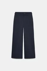 Pantalón negro con finas rayas verticales blancas, de tiro medio y corte ancho. Presenta bolsillos delanteros y traseros, cintura doble tipo bóxer con botones y cierre frontal con cremallera y ganchos metálicos. Detalle de franja marrón en los laterales.