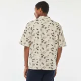Camisa de manga corta color beige con estampado de palmeras y motivos playeros en negro.