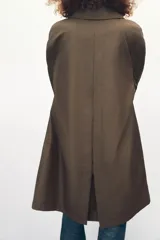 Trench color marrón con cuello solapa y manga tres cuartos. Tiene bolsillos delanteros y cierre frontal con cremallera y botones ocultos por solapa.