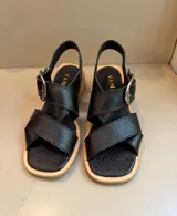 Sandalia de cuero vacuno color marrón, con tiras cruzadas en el empeine y hebilla metálica plateada. Posee taco de madera de 5 cm de altura y horma de punta cuadrada.