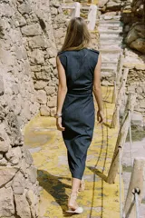 Vestido midi de denim azul oscuro, con cuello redondo y sin mangas. Presenta detalle de frunces en la cintura, cinturón elástico con hebilla dorada y cierre lateral con cremallera oculta. Tiene falsos botones delanteros.